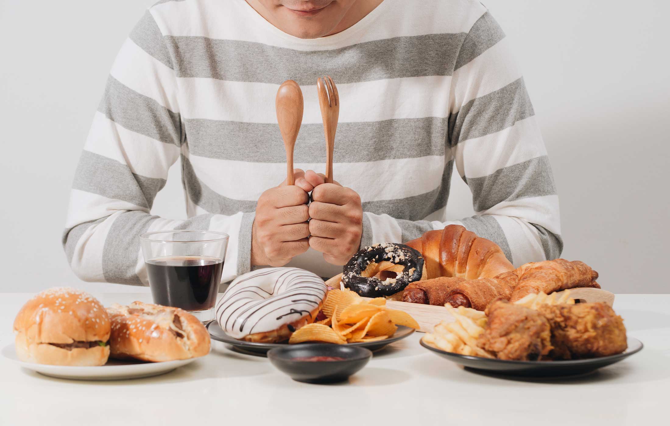 Faceless person sitting at table with donuts, hamburgers, hot dogs, chips, pastries and fried chicken